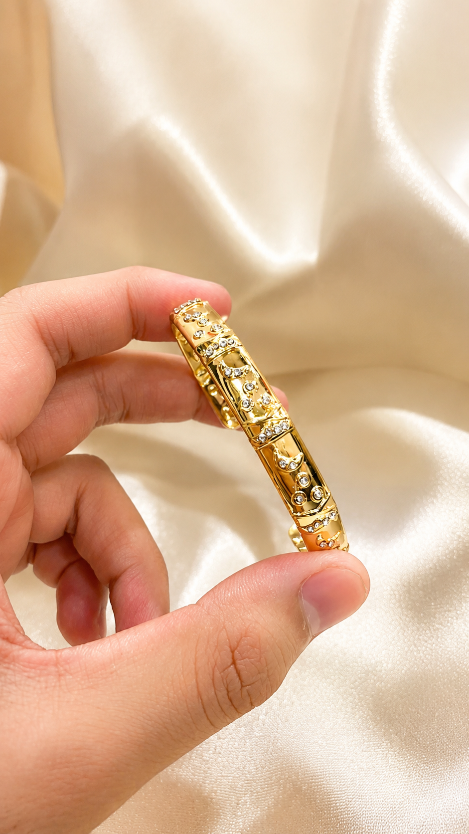 A hand holding a gold-plated bangle bracelet decorated with small clear crystals, set against a draped cream satin fabric background.