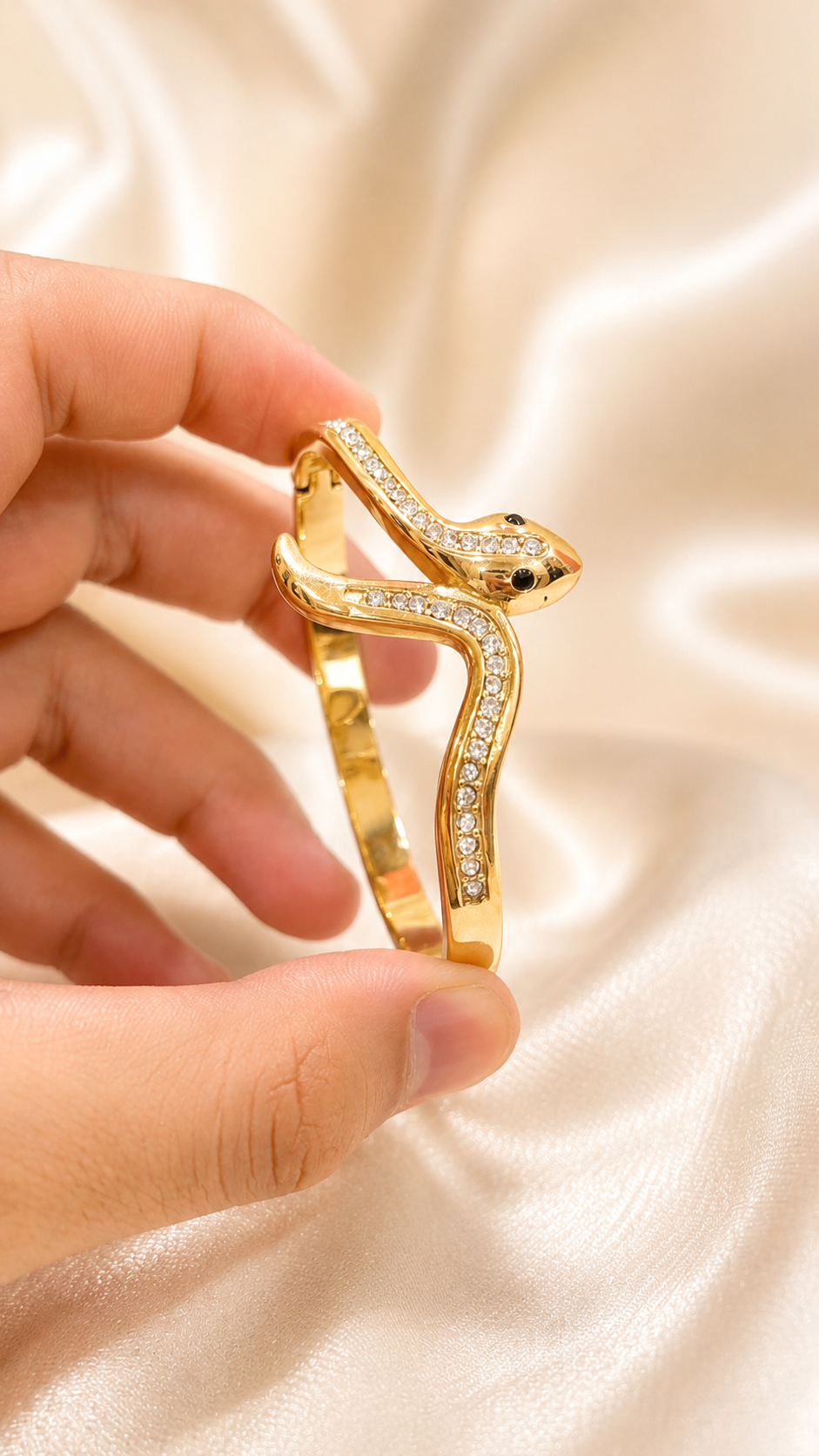 A gold snake-shaped bangle bracelet with pavé-set clear stones, held by a hand against a draped cream satin fabric background.