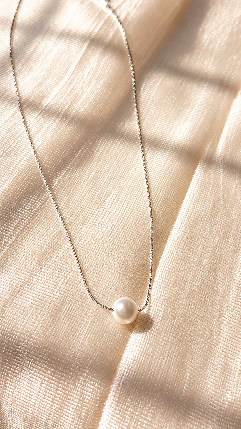 A delicate silver chain necklace with a single white pearl pendant, laid out on a textured beige fabric with soft window shadow overlays.
