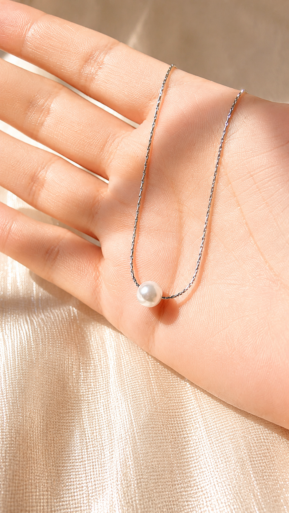 A delicate silver chain necklace with a single white pearl pendant, draped across an open hand against a soft beige fabric background.