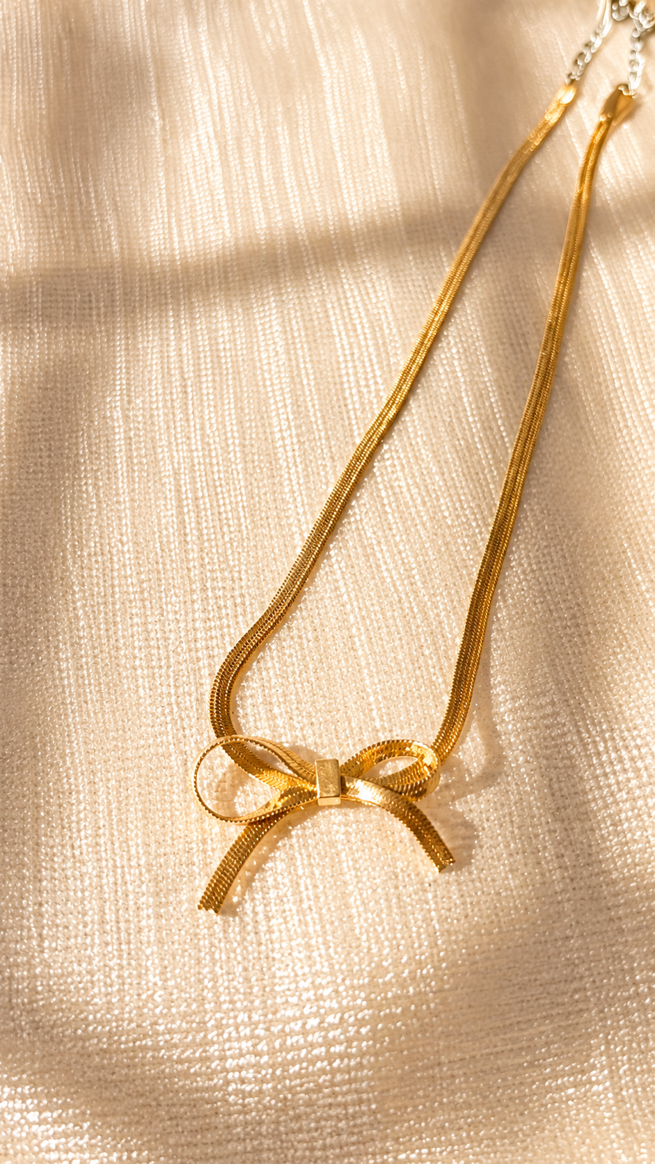 A delicate gold snake chain necklace featuring a small bow pendant, laid flat on a textured cream-colored fabric with soft, natural shadows.
