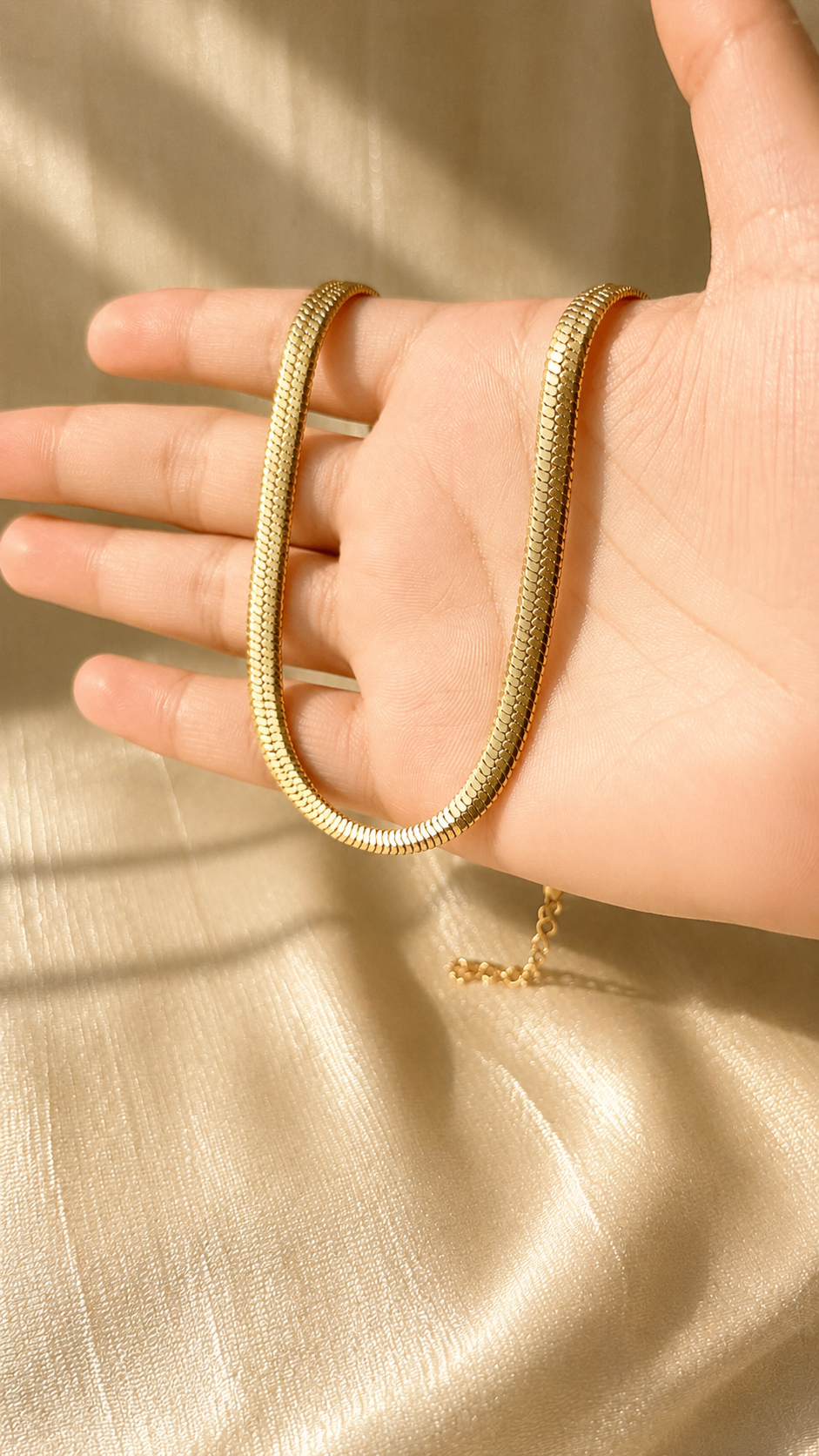 A gold snake chain bracelet draped over a person's hand, set against a soft beige satin fabric background with warm sunlight and shadows.