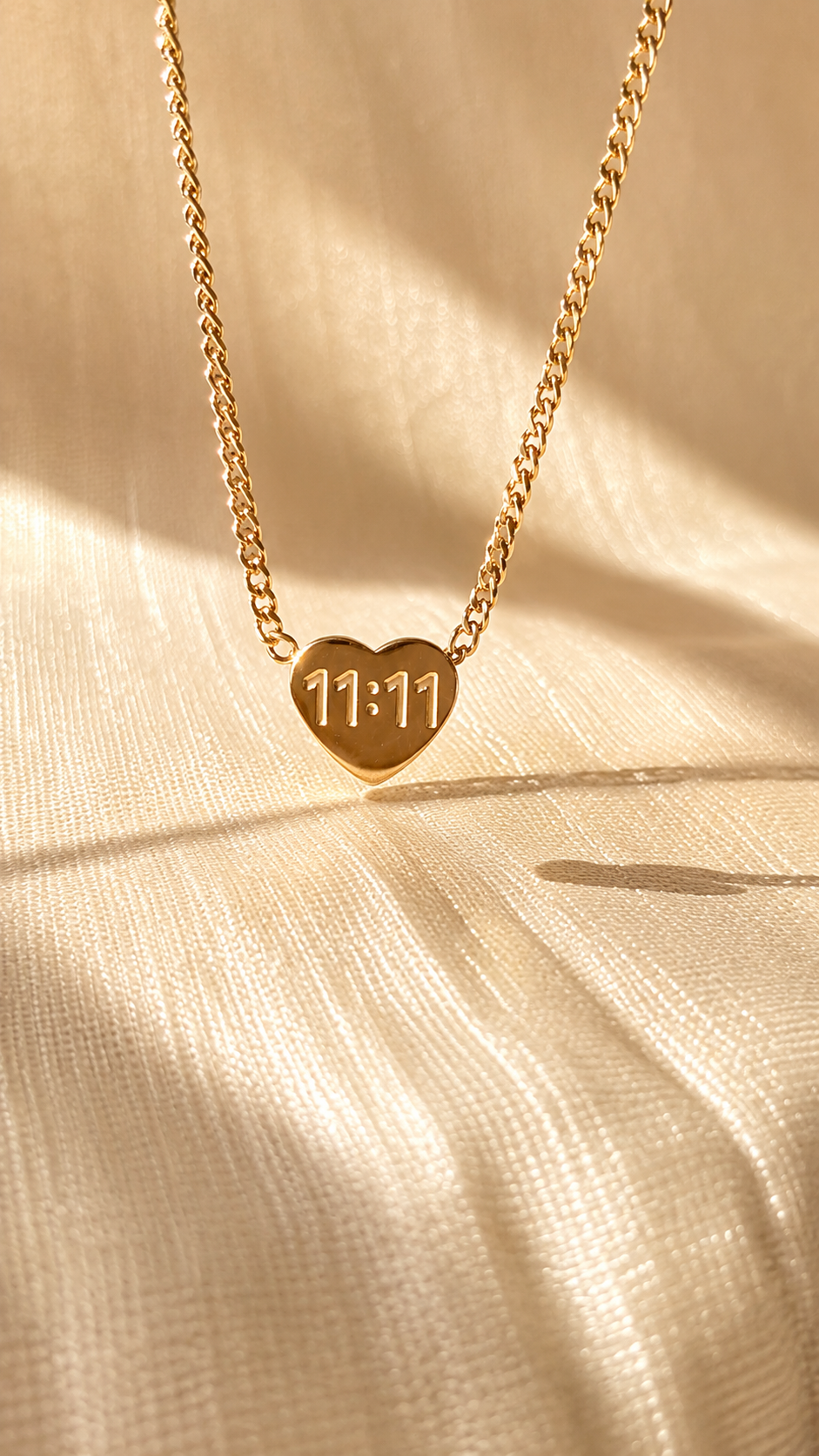 A gold heart-shaped pendant necklace engraved with '11:11' on a matching chain, displayed on draped beige fabric with soft shadows.